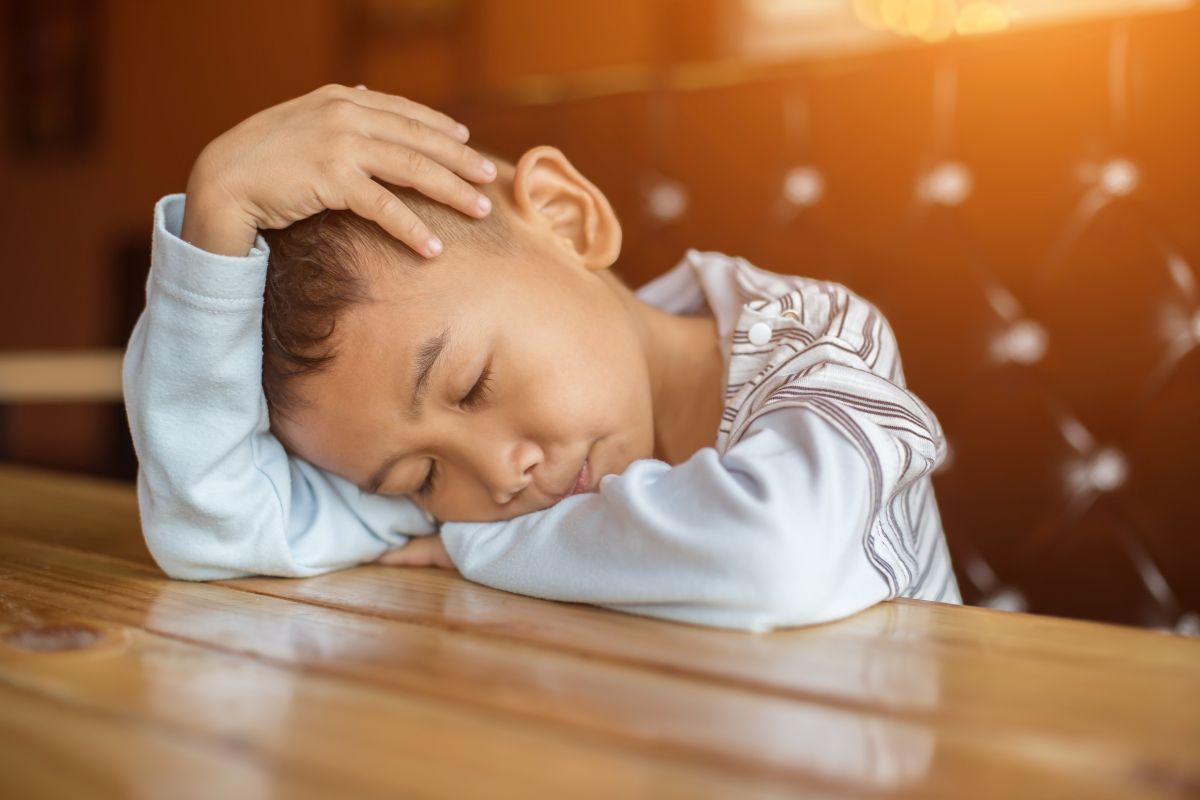 Young boy with eyes closed rests his head on folded arm on a wooden table, sleeping peacefully in a warm-lit setting.